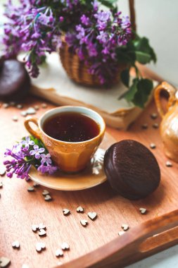 Yeşillik taze çiçeklerle ev yapımı çay partisi. Siyah çayla altın porselen fincan, çikolatalı pasta, mor leylak çiçekleri hasır sepetin içinde kalplerle süslenmiş ahşap tepside. dikey
