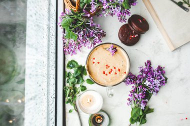 Köpüklü dalgona kahvesi, çikolatalı kurabiyeler, yanan mumlar ve ıslak pencerede sulu mor leylak çiçekleri yağmurdan damlayan damlalar. Sıcak, güzel kahvaltı, kötü yağmurlu hava manzarası.