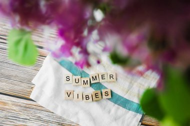 Tahta bir masanın üzerinde beyaz bir pamuk havlunun üzerindeki tahta kare harflerden oluşan yazlık titreşimler. Üzerinde mor, yemyeşil bir demet, üst görünüm, blog için hazır yüksek kaliteli içerik.