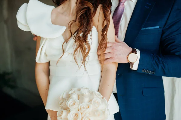 Stylish wedding picture. The groom in a blue suit gently embraces the bride in a white stylish dress made of stiff fabric. Puffy wedding dress sleeve, flounce sleeve on an unusual wedding dress.