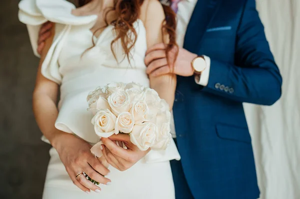 花嫁の手に新鮮な白いバラの結婚式の花束 スタイリッシュな結婚式の写真 ブルーのスーツを着た新郎は 花嫁を優しく包み込みます白いスタイリッシュなドレスで硬い生地で作られていますパフィーフロンススリーブ — ストック写真
