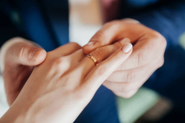 The groom in a blue suit puts a gold ring on the hand of the bride in a white stylish dress. Hands of the newlywed close-up, no faces.