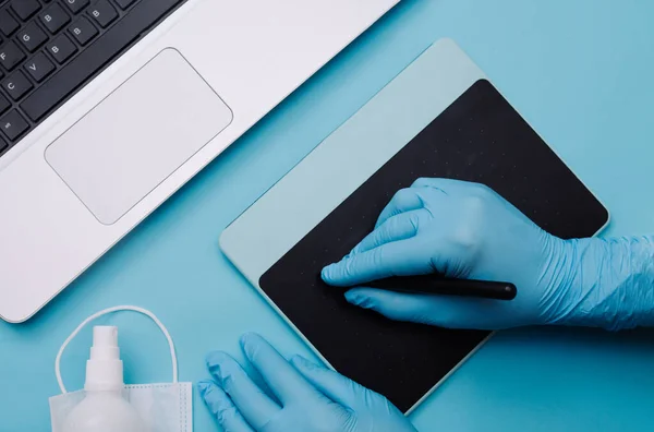 A hand in a blue medical surgical protective glove draws with a stylus on a graphic planchette next to a laptop on a clear blue background. Home office in Coronovirus quarantine mode
