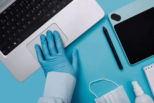 A hand in a blue medical surgical protective glove draws works on a laptop on a clear blue background. Home office with a graphic tablet, stylus, mask and sanitizer in Coronovirus quarantine mode