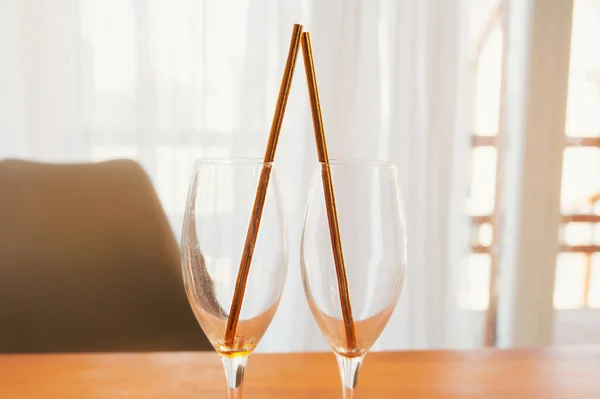 Two beautiful champagne glasses with gold paper tubes on a wooden table against the background of a large window with light curtains. Romantic date for two at home
