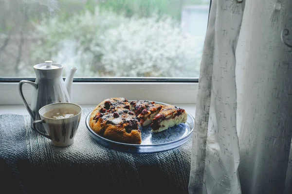 cozy home tea party by window in bad weather. Porcelain white teapot and cup with tea, homemade berry pie on a round blue plate against a flowering tree on the street outside the window with curtain