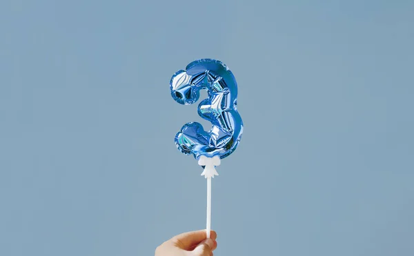 El ele tutuşan mavi folyo helyum 3 numaralı balonu mavi arka planda beyaz bir plastik çubukta şişiriyor. Parlak bayram selamlama düzeni