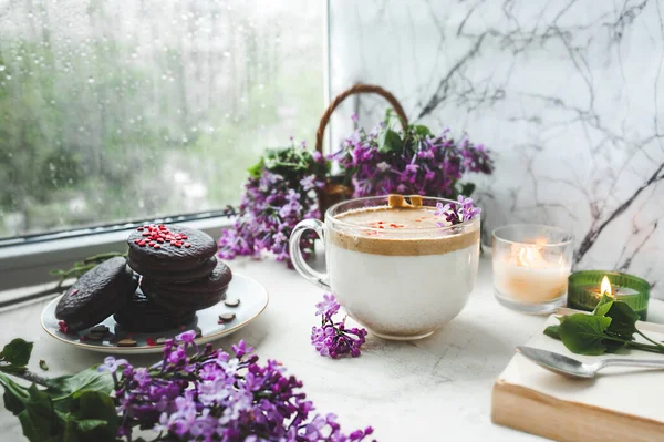 dalgona coffee with foam, chocolate cookies, burning candles and fresh lush purple-lilac flowers at the wet window with drops and drips from the rain. cozy beautiful Breakfast in bad wet weather