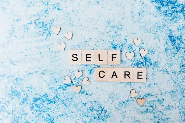 self care lettering in square wooden letters with small wooden hearts on a blue and grey concrete background. layout about Taking care of yourself and your health