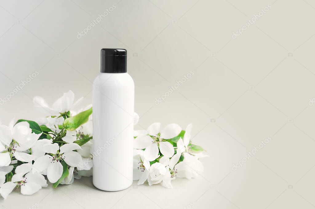 white clean bottle with a black cap with a blossoming white sprig of lush flowers with green leaves on white background. Cosmetic layout. Skin and body care mock up