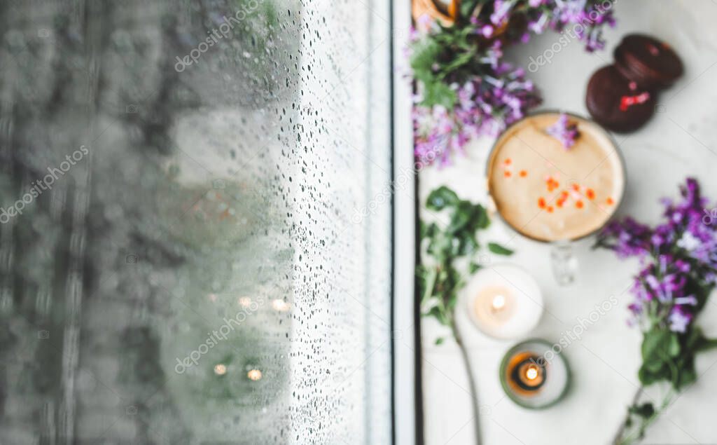 raindrops on the window glass against the background of blurred coffee dalgon with foam, chocolate cookies, burning candles and fresh lush purple-lilac flowers, selective focus, art photo