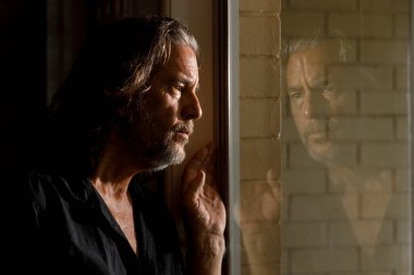 Kendini izole eden, evde kalan, tuğla evin camındaki yansımasını düşünen, pencereden dışarı bakan uzun saçlı, sakallı, akıl sağlığı yerinde bir adam.