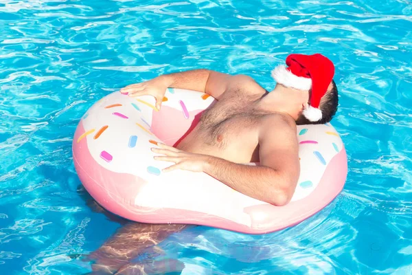 top view of young drunk guy in santa claus hat swim with pink circle in pool. drunk guy on vacation in the hotel.