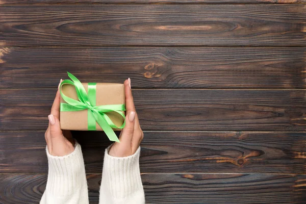 Woman hands give wrapped valentine or other holiday handmade present in paper with red ribbon. Present box, decoration of gift on white wooden table, top view with copy space.