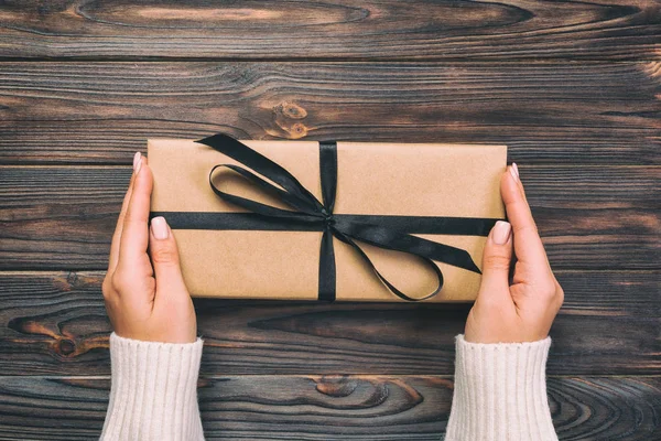 Woman hands give wrapped valentine or other holiday handmade present in paper with black ribbon. Present box, decoration of gift on dark wooden table, vintage top view with copy space.