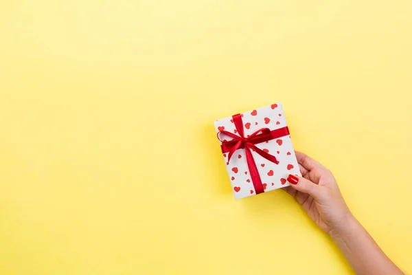 Woman hands give wrapped valentine or other holiday handmade present in paper with red ribbon. Present box, red heart decoration of gift on yellow table, top view with copy space for you design.