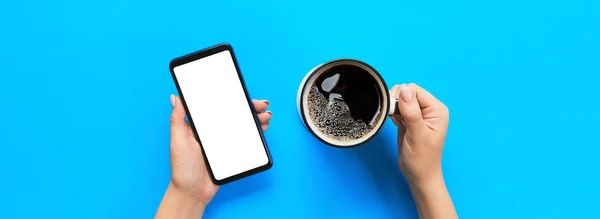 Female hands holding black mobile phone with blank white screen and mug of coffee. Mockup image with copy space for you design. Top view banner on blue background, flat lay.