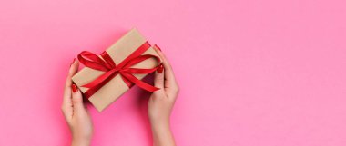 Kadın elleri, paketlenmiş sevgililer günü hediyesi olarak kırmızı kurdeleli el yapımı hediyeler verir. Hediye kutusu, hediye süslemesi pembe masada, En üstteki afiş fotokopi alanı sizin için tasarlandı.