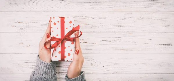 Woman hands give wrapped valentine or other holiday handmade present in paper with red ribbon. Present box, red heart decoration of gift on wooden table, top view with copy space Toned web banner.