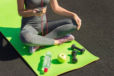 Oturma ve spor salonunda smartphone kullanarak genç fitness sporcumuz. Yoga mat müzik su, halter ve ealthy elma dinler botte ile kız. Spor ve sağlıklı kavramı. Üstten Görünüm.