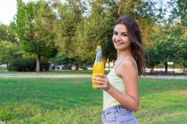 Genç kız sıcak bir günde elinde bir şişe soğuk meyve suyu ya da limonata tutarak parkta yürüyor.