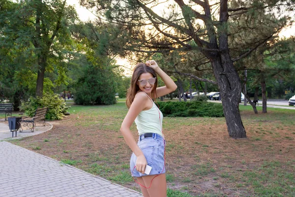 Giovane ragazza carina elegante sta ascoltando musica in cuffia sul telefono cellulare e ballare nel parco. Bella donna è rilassante e divertirsi — Foto Stock