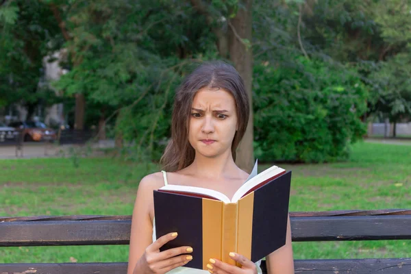 Confused and displeased girl is reading a book on the bench in the park — Stock Photo, Image