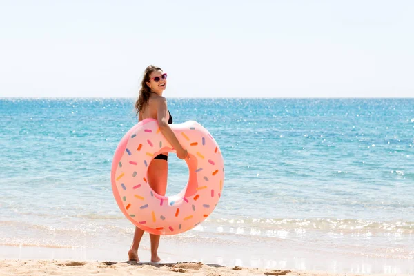 Сексуальная симпатичная счастливая женщина бегает по пляжу с розовым резиновым накладным кольцом в руке. Летние каникулы и отдых — стоковое фото
