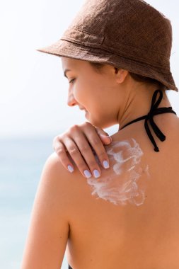 Şapkalı genç kız cildini korumak için sırtına güneş kremi sürüyor.