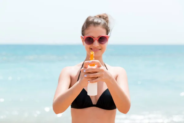 Młoda kobieta trzyma spray kremu do opalania na ciało przed kamerą na plaży — Zdjęcie stockowe