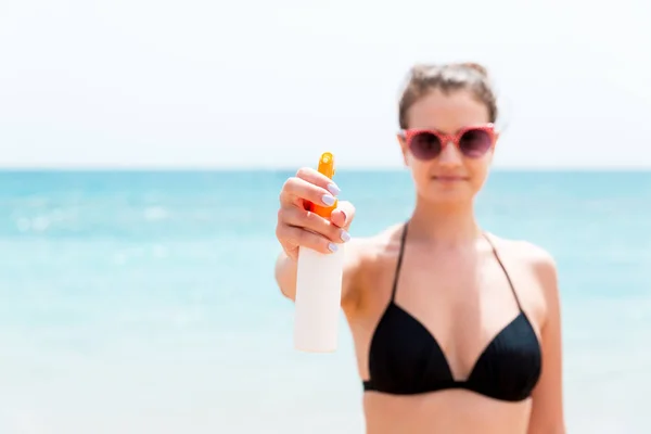 Młoda kobieta trzyma spray kremu do opalania na ciało przed kamerą na plaży — Zdjęcie stockowe