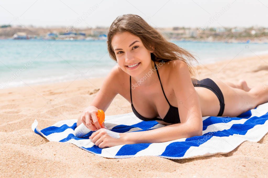 Young happy woman is resting on the towel near the sea and protects her skin on the hand with sunscreen from the spray