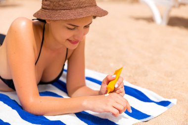 Güzel bayan kumsalda kumların üzerinde güneşleniyor ve ellerini güneş kremiyle koruyor.