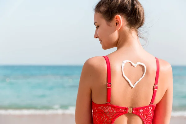 Bella ragazza in costume da bagno rosso ha la crema solare a forma di cuore sul retro in spiaggia — Foto Stock