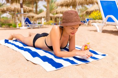 Güzel bayan kumsalda kumların üzerinde güneşleniyor ve ellerini güneş kremiyle koruyor.