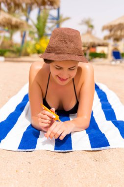 Gülümseyen genç kadın kumsaldaki kumların üzerinde çizgili havluya uzanmış ve eline güneş kremi sürüyor.