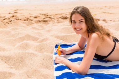 Çekici kadın sahilde çizgili bir havlu üzerinde yatıyor ve elinde bir tüp güneş kremi sıkma