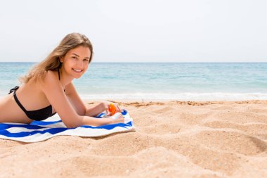 Genç mutlu kadın denizin yanındaki havluda dinleniyor ve cildini güneş kremiyle spreyden koruyor.
