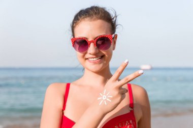 Genç bir kadın barış jesti yapıyor ve elinde kumsalda güneş şeklinde bir şey var.