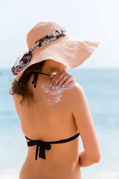 Hermosa mujer con sombrero está poniendo crema solar en su hombro en la playa — Foto de Stock