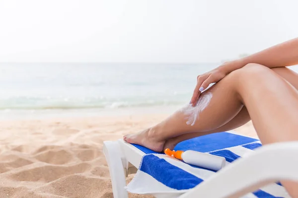 Mujer joven está descansando en la tumbona en la playa y protege su piel aplicando bloqueador solar en su pierna — Foto de Stock