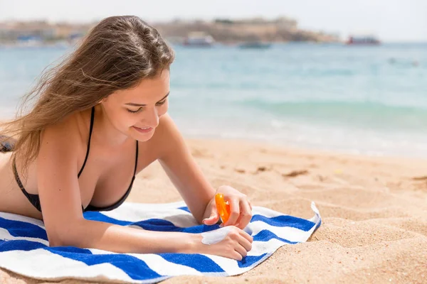 Sevimli ve gülümseyen bir kadın eline güneş kremi sürüyor. Kumsalda havluyla güneşleniyor.