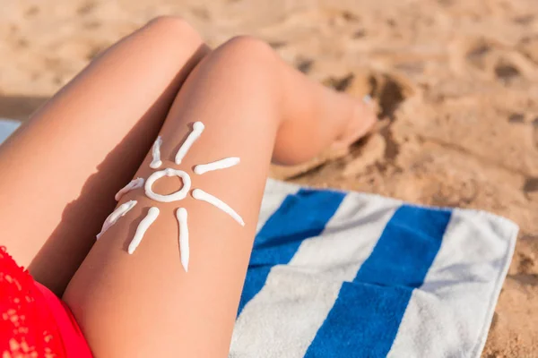Creme solar é desenhado em forma de sol na perna da mulher bronzeada que está relaxando na toalha na praia. Conceito de cuidados oncológicos — Fotografia de Stock