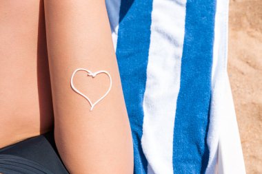 Deniz tarafından havlu üzerinde güneşlenen kadının bacağında kalp şeklinde güneş kremi