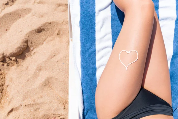 Αντηλιακό με τη μορφή καρδιάς στο πόδι της γυναίκας που κάνει ηλιοθεραπεία στην πετσέτα δίπλα στη θάλασσα — Φωτογραφία Αρχείου