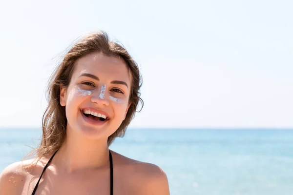 Beautiful young woman is posing at the camera with suncream on her face over the sea background — Stock Photo, Image