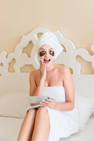 surprised or shocked young woman with under eye patches got a bad news and using mobile phone in bathrobe lying in bed. Happy girl taking care of herself. Beauty skincare and wellness morning concept