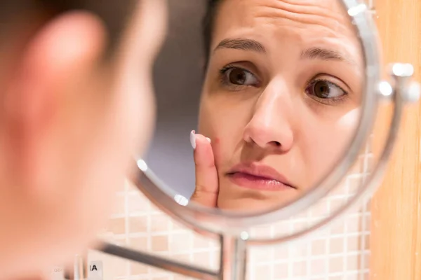 Young girl in front of a bathroom mirror putting cream on a red pimple. Beauty skincare and wellness morning concept