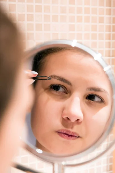 Beautiful woman with tweezers is plucking eyebrows while looking — Stock Photo, Image