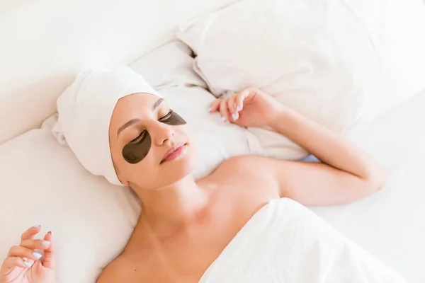 Hermosa mujer joven con parches debajo de los ojos en albornoz acostado en la cama. Chica feliz cuidando de sí misma. Belleza cuidado de la piel y bienestar concepto de la mañana — Foto de Stock